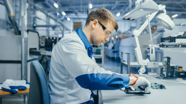 istock Young Man in Blue and White Work Coat is Using Plier to Assemble Printed Circuit Board for Smartphone. Electronics Factory Workers in a High Tech Factory Facility. 1145276382