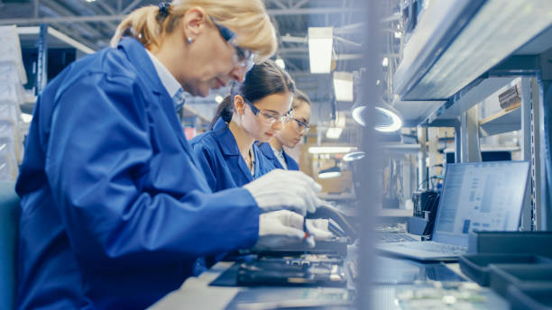 istock Female Electronics Factory Worker in Blue Work Coat and Protective Glasses is Assembling Smartphones with Screwdriver. High Tech Factory Facility with more Employees in the Background. 1145276393