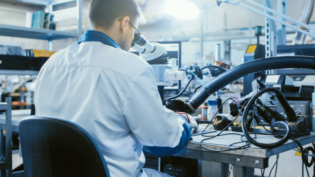 istock Back View Shot of an Electronics Factory Worker in White Work Coat Inspects a Printed Circuit Board Through a Digital Microscope. High Tech Factory Facility. 1145276398
