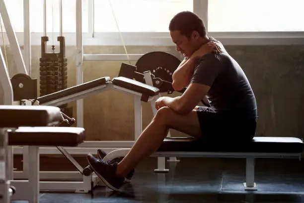 Photo of asian Muscular man suffering from shoulder injury while exercising at gym in morning light . workout , exercises , bodybuilder training , sport guy rubbing his back and neck pain .  backache