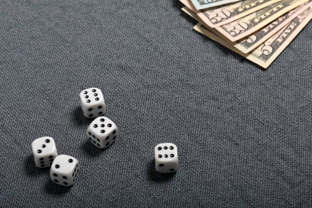 istock White dice with black markings. They lie on a surface covered with a coarse gray cloth. Nearby are a few dollar bills of different denominations. 1145292321