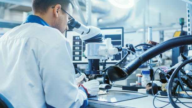 istock Electronics Factory Worker in White Work Coat is Soldering a Printed Circuit Board Through a Digital Microscope. High Tech Factory Facility. 1145292407