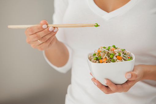 Slim young woman (torso) holding a chinese-like healthy food bowl in one han, and chopsticks in the other. High key lighting.