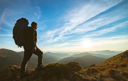 The man standing with a camping backpack on the rock with a picturesque sunset