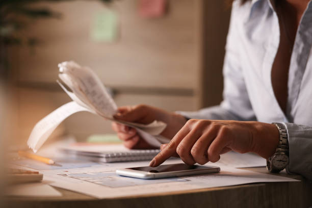 Close up of businesswoman using smart phone while calculating her bills in the office. Close up of businesswoman going though finances and using mobile phone as calculator. freelance work stock pictures, royalty-free photos & images