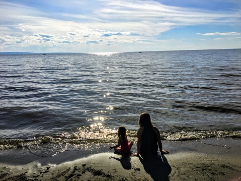A young mother and her toddler daughter sitting together alone on a sandy beach watching the sparkling lake water of lesser salve lake Alberta, Canada. It is nearing sundown and their backs are turned