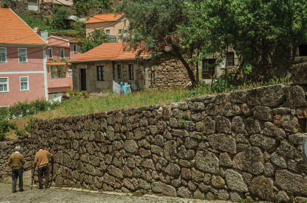 Two old people walking down the alley on slope next to stone wall, in a sunny day at Alvoco da Serra. A cute village clinging on a steep valley in the Serra da Estrela highland on eastern Portugal.