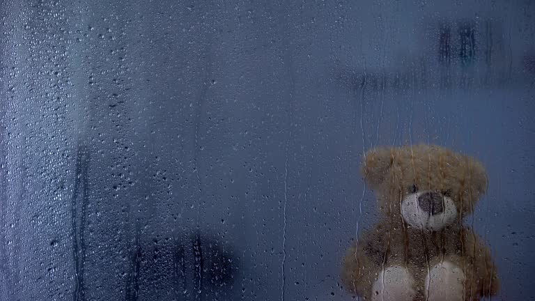 Teddy bear on sill behind rainy window in orphanage, hope and support symbol