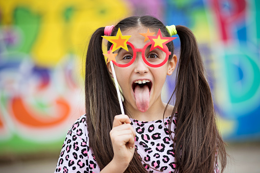 Fun young girl sticking out her tongue as she holds a party accessory of colorful glasses with stars to her eyes against a multicolored background