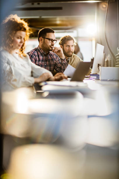 Group of colleagues working on desktop PC in the office. Group of colleagues working at corporate office. Focus is on pensive man with glasses working on PC with his colleague. Copy space. young graphic designer stock pictures, royalty-free photos & images