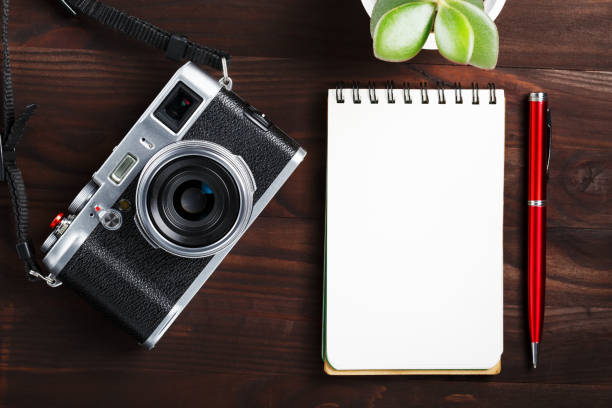 istock Classic camera with blank notepad page and red pen on dark brown wooden table, vintage table with telephone and green flower 1145396928