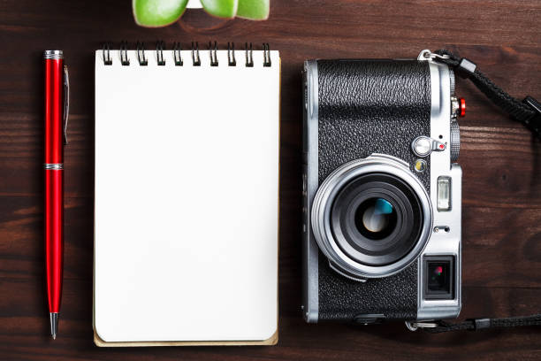 istock Classic camera with blank notepad page and red pen on dark brown wooden table, vintage table with telephone and green flower 1145397720