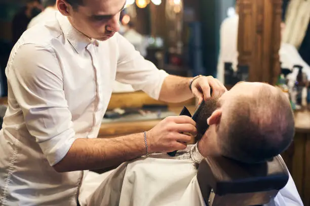 Photo of Male client with beard sitting in hairdresser chair. Serious man with long brown beard. Modern popular lumberjack style.