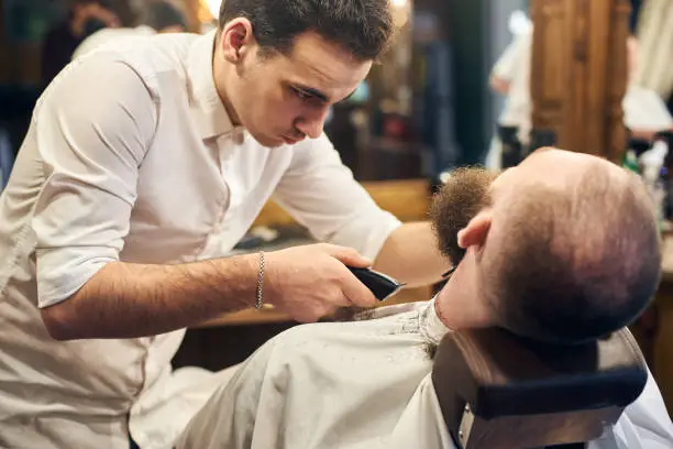 Photo of Male client with beard sitting in hairdresser chair. Serious man with long brown beard. Modern popular lumberjack style.