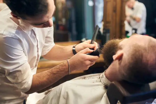 Photo of Male client with beard sitting in hairdresser chair. Serious man with long brown beard. Modern popular lumberjack style.