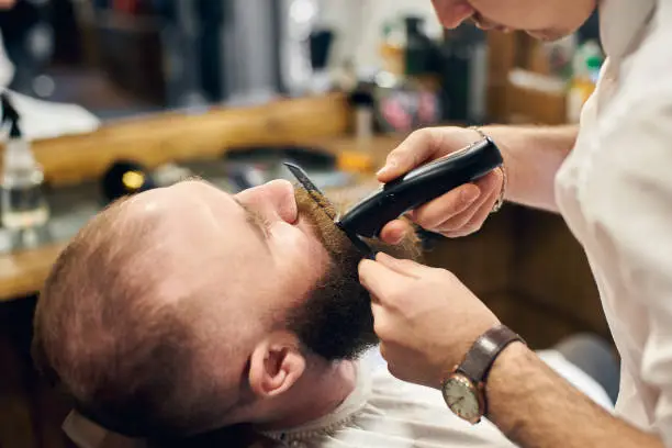 Photo of Male client with beard sitting in hairdresser chair. Serious man with long brown beard. Modern popular lumberjack style.