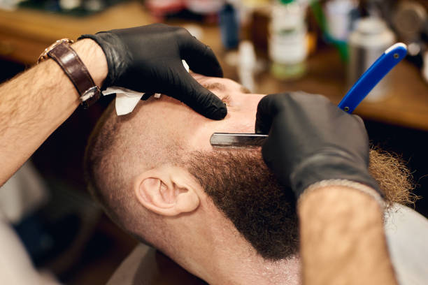 istock Bearded client sitting in chair for shaving by barber hands with sharp blade. Using traditional tools for male face care 1145397836