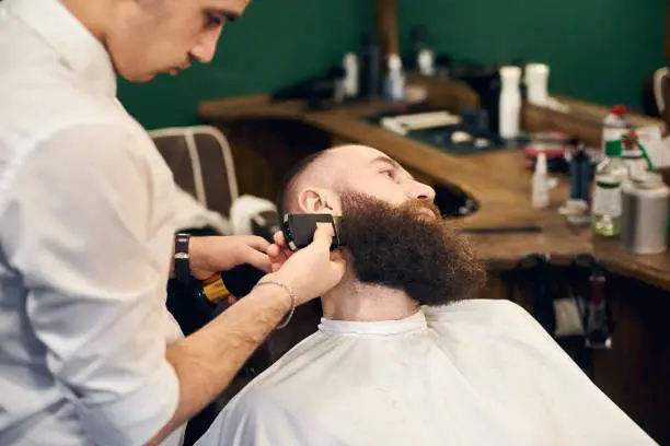 Photo of Male client with beard sitting in hairdresser chair. Serious man with long brown beard. Modern popular lumberjack style.