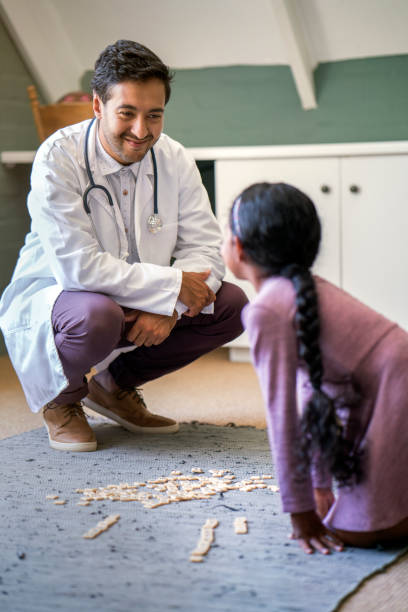 Smiling male doctor visiting girl at home Smiling male pediatrician looking at patient in room. Girl is playing with healthcare worker. Doctor is visiting on house call. squatting position photos stock pictures, royalty-free photos & images