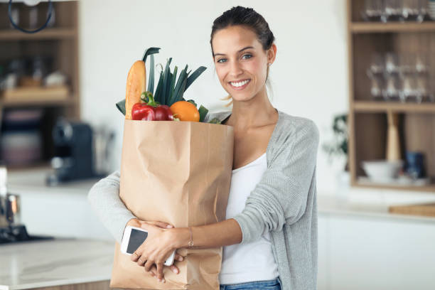 台所で新鮮な野菜とショッピングバッグを保持しながら、カメラを見ている美しい若い女性。 - paper bag groceries food vegetable ストックフォトと画像