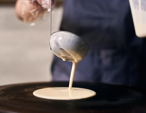 Woman chef in blue apron pour portion of liquid dough by hands in gloves with ladle on electric cooktop for baking pancakes, crepes. Cheese and tomato slice in plate on blurred background. Front view.