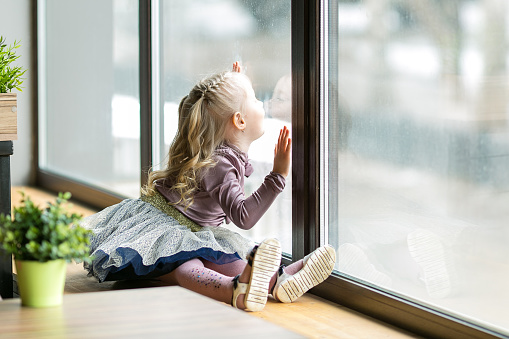 A little girl, 4-5 years old  is sitting on the windowsill of a large window in a cafe.