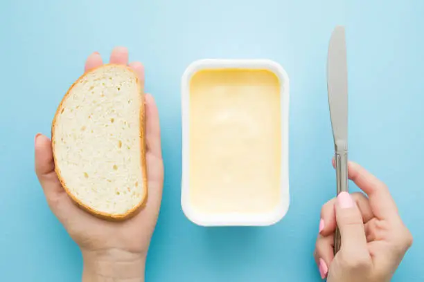 Woman's hands holding slice of white bread and knife. Opened plastic pack of light yellow margarine on pastel blue desk. Preparing breakfast. Point of view shot. Closeup. Top view.