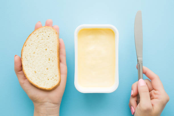 woman's hands holding slice of white bread and knife. opened plastic pack of light yellow margarine on pastel blue desk. preparing breakfast. point of view shot. closeup. top view. - margarine dairy product butter close up imagens e fotografias de stock