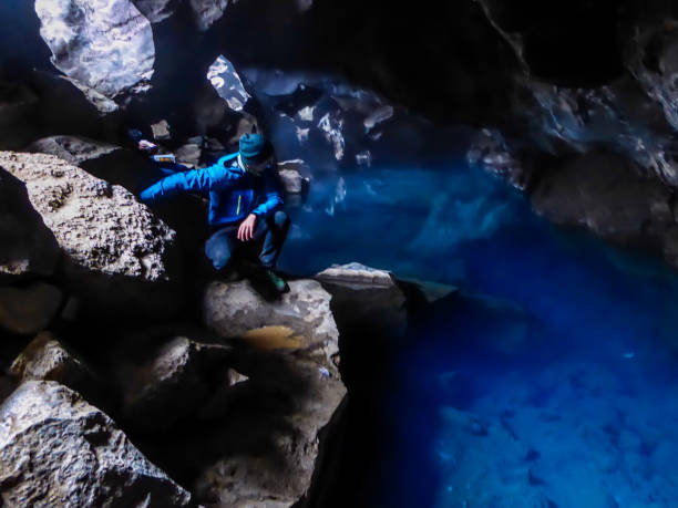 Iceland - Young man at the Grjótagjá Cave with extremely bue water A young man wearing blue jacket squats at the rock in  Grjótagjá Cave, looking down at the crystal blue water. Famous Game of thrones location. The water in this cave is full of minerals, it's hot pot grjótagjá thermal spring stock pictures, royalty-free photos & images
