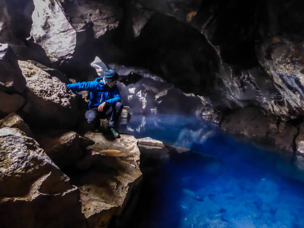 Iceland - Young man at the Grjótagjá Cave with extremely bue water A young man wearing blue jacket squats at the rock in  Grjótagjá Cave, looking down at the crystal blue water. Famous Game of thrones location. The water in this cave is full of minerals, it's hot pot grjótagjá thermal spring stock pictures, royalty-free photos & images