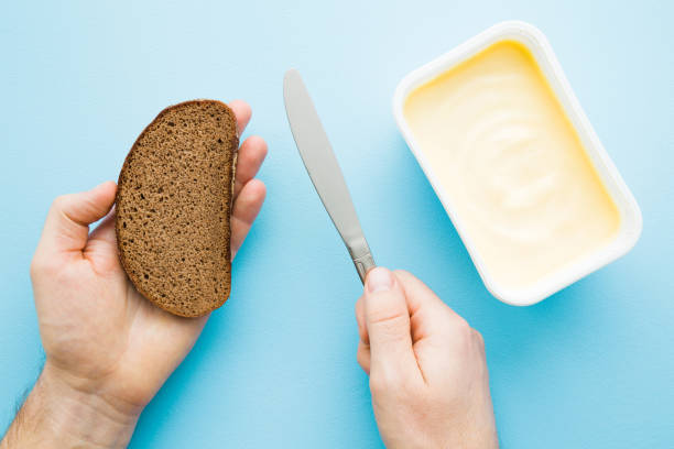 man's hands holding slice of brown bread and knife. opened plastic pack of light yellow margarine on pastel blue desk. preparing breakfast. point of view shot. closeup. top view. - margarine dairy product butter close up imagens e fotografias de stock