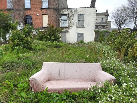 Abandoned couch in derelict house's overgrown front garden with the property's windows boarded up with cement bricks.