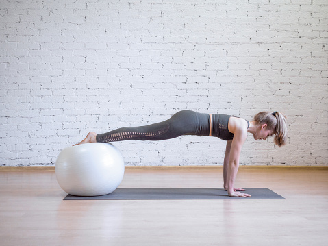 Caucasian woman doing plank position using math and fitness ball, loft background, toned blue. Yoga, pilates, fitness, wellness, balance and smart body concept