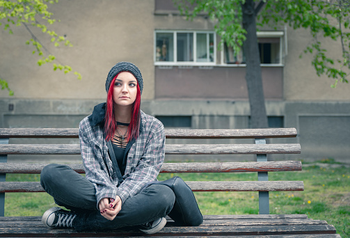 Young beautiful red hair girl sitting alone outdoors on the wooden bench on the street with hat and shirt feeling anxious and depressed after she became a homeless person