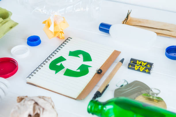 istock Recycle paper notebook with Green eco symbol surrounded with non recycling garbage and reusable waste as metal, plastic, old paper, glass products on the white wooden background. Selective focus. 1145481377