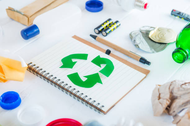 istock Recycle paper notebook with Green eco symbol surrounded with non recycling garbage and reusable waste as metal, plastic, old paper, glass products on the white wooden background. Selective focus. 1145481380