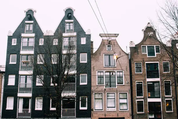 Photo of Traditional bricked Dutch houses in Amsterdam, Netherlands. Narrow black and pastel facade walls with tall windows against gray cloudy sky.