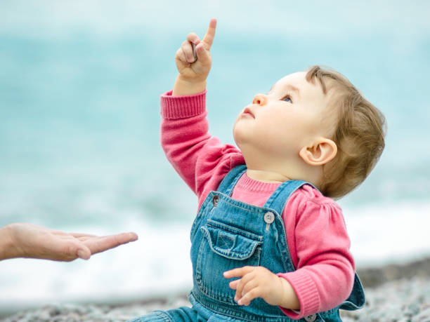 Baby in denim sits on the beach and looks up Baby and mother playing with pebbles. One-year-old child on the beach in spring close-up. Little cute baby in denim sits and looks up. Playful toddler in points his finger emotionally. baby1 stock pictures, royalty-free photos & images