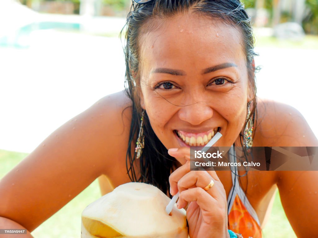 lifestyle natural portrait of 30s or 40s happy and attractive Asian Indonesian woman in bikini drinking coconut water at tropical resort swimming pool smiling cheerful and relaxed enjoying holidays Only Women Stock Photo