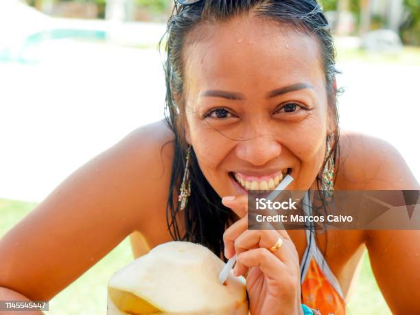 Lifestyle Natural Portrait Of 30s Or 40s Happy And Attractive Asian Indonesian Woman In Bikini Drinking Coconut Water At Tropical Resort Swimming Pool Smiling Cheerful And Relaxed Enjoying Holidays Stock Photo - Download Image Now