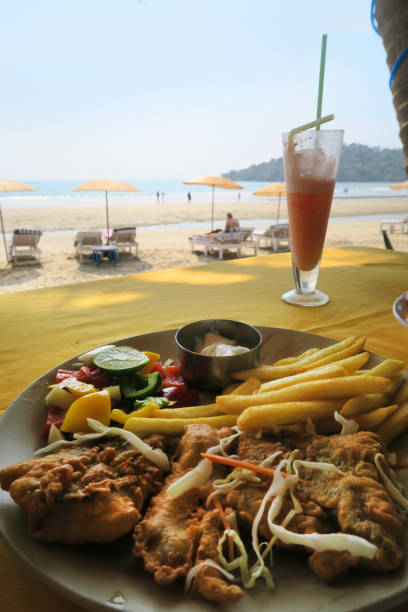 Image of Indian beach fish and chips with battered kingfish fish (not cod) and French fries, cocktail drink under palm tree, salad, homemade mayonnaise, sun loungers, sea, bean bags in background, seafood restaurant dining on Palolem beach, Goa, India Stock photo of Indian beach fish and chips (fish supper), which comprises pieces of battered kingfish fish (not traditional cod and chips) as well as crispy French fries. This seafood restaurant is especially popular with tourists on vacation and holidaymakers on holiday in Goa, since it sits directly on the beach, next to the sand and the distant sea, as well as the sunloungers and beach thatched parasols. The cod and chips meal is accompanied by a fruit juice based cocktail drink under a coconut palm tree, and has been served with salad, a dish of mayonnaise, next to the bean bags and overlooking Palolem beach, Goa, South India. Of note, further beaches lie to the south of Goa, in the neighbouring state of Kerala. palolem beach stock pictures, royalty-free photos & images