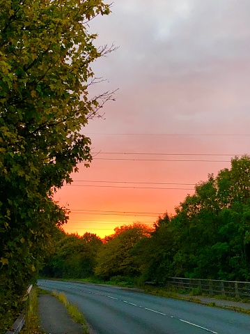 Stock photo showing an early morning sunrise on a peaceful English road / highway. This tarmac road is crossing over a railway bridge, while the focus of the photo is the bridge pink orange sunrise / sunset in the background. Above the morning sunrise and quiet traffic free road are grey stormy clouds blowing across overhead, while green trees and hedgerows edge the road and pavement sidewalks.