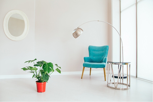 Conceptual light  interior of living room with blue armchair, metallic standing lamp,  glass round coffee table , oval mirror on white wall, monstera plant in red vase on the floor and large window. Concept of minimalizm.