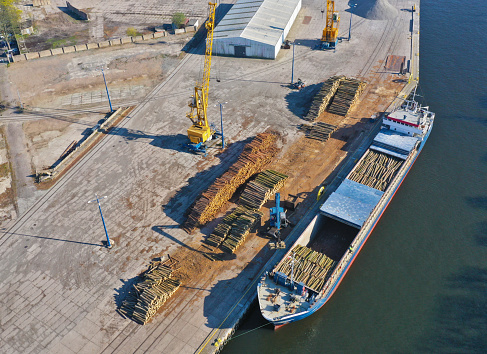 Aerial view on cargo ship in port bank loading wood stack on board using crane.