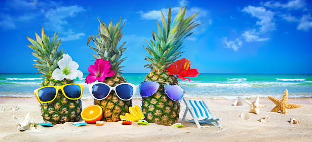 Attractive pineapples in stylish sunglasses on the sand beach against turquoise sea. Tropical summer vacation concept