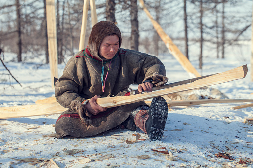 Residents of the far north,  the pasture of Nenets people, the dwelling of the peoples of the north of Yamal, a man makes wooden sleds by hand with a an ax
