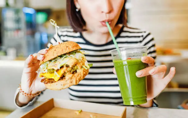 Photo of A young woman drinks green smoothies and eats a burger in a vegan fast food restaurant