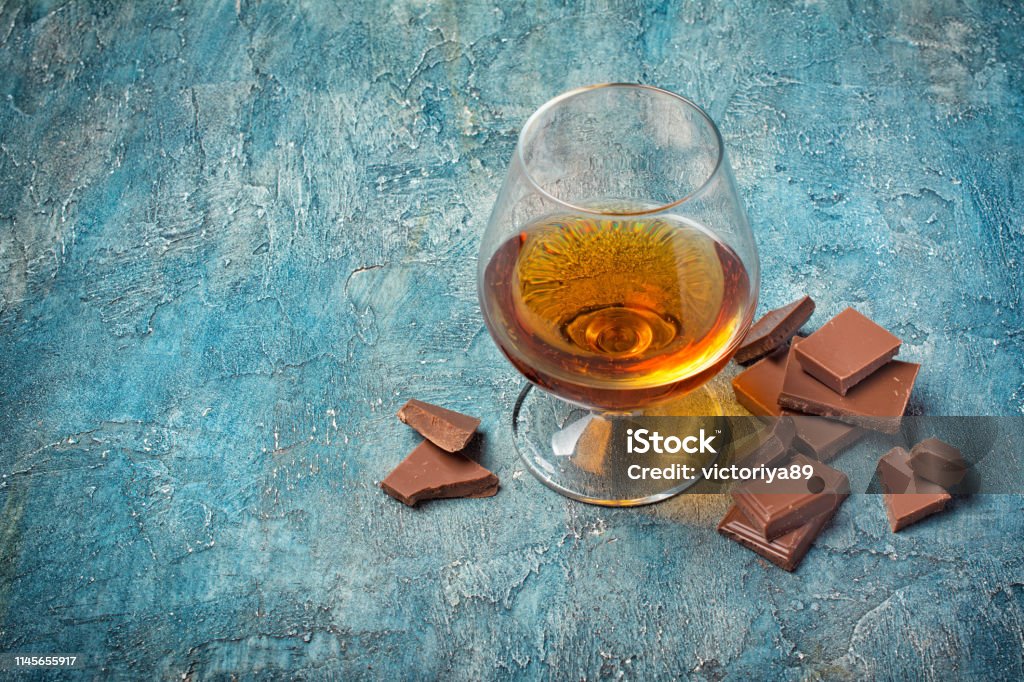 Strong alcoholic beverage cognac in sniffer glass with chocolate bar Strong alcoholic beverage cognac in sniffer glass with chocolate bar for tasting on blue concrete background with copy space Addiction Stock Photo