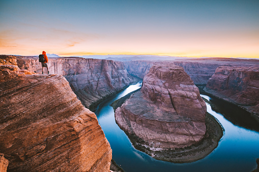 A male hiker is standing on steep cliffs enjoying the beautiful view of Colorado river flowing at famous Horseshoe Bend overlook in beautiful post sunset twilight on a summer evening, Arizona, USA