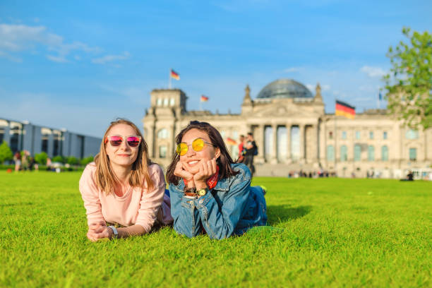 dos jóvenes chicas felices que llevan gafas de sol tumbados sobre una hierba y se divierten frente al edificio bundestag en berlín. estudiar en el extranjero y viajar en alemania concepto - berlin germany fotografías e imágenes de stock